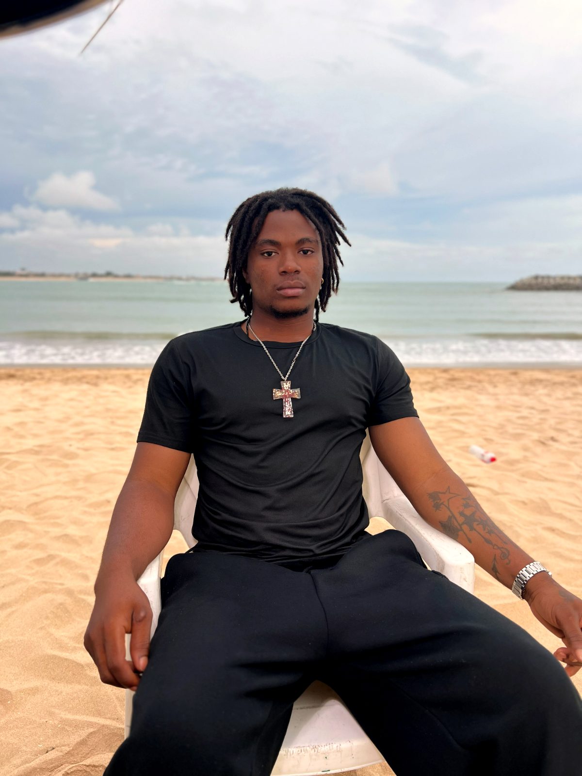 OIM THUG assis face à la mer, t-shirt noir et croix