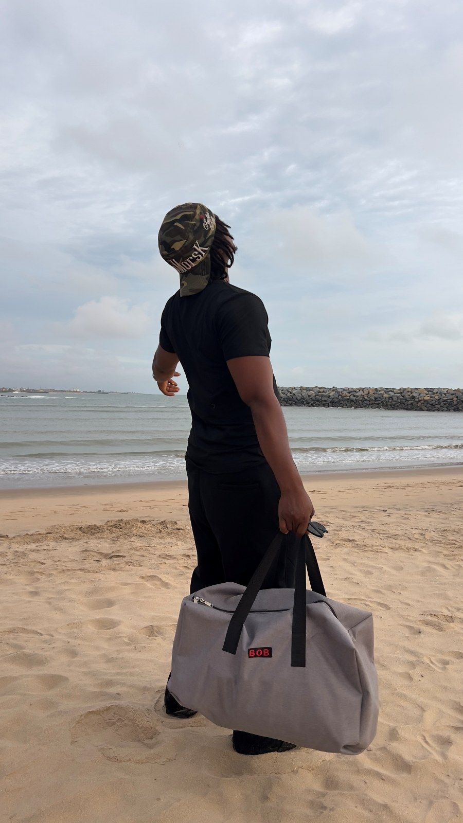 OIM THUG sur la plage, un sac à la main, le bras tendu vers l’horizon